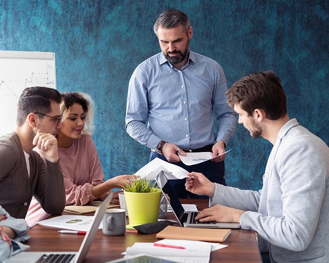 Five people gathered around a wooden table with laptops and documents, with one person standing and presenting near a flip chart.