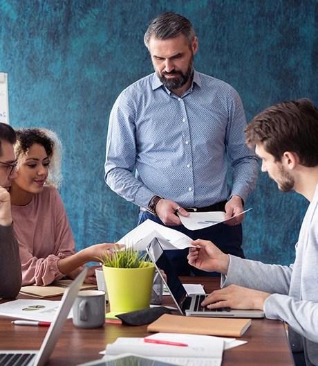 Five people gathered around a wooden table with laptops and documents, with one person standing and presenting near a flip chart.