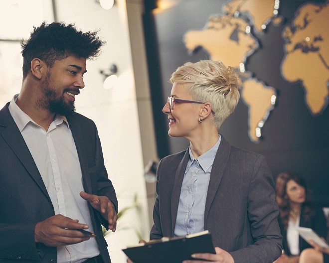 Two individuals in business attire talking in front of a wall-mounted world map with illuminated markers, one holding a clipboard and the other gesturing.