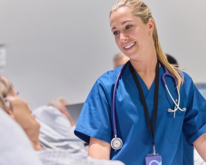 Healthcare professional in blue scrubs and a stethoscope smiling while speaking to a patient in a hospital bed, with other patients visible in the background.