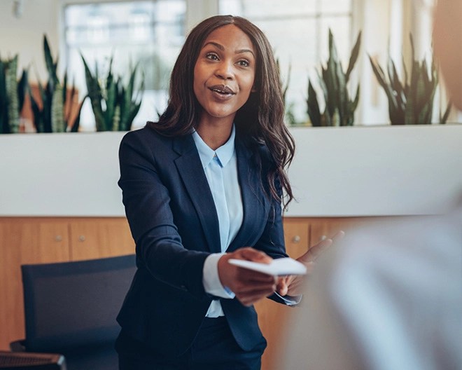 Person in a dark suit and light blue shirt holding a white card while speaking to another individual in a modern office with green plants and wood paneling.