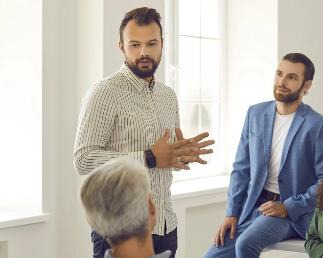 Group of people in a well-lit room with large windows, with one person standing and gesturing while others sit and listen attentively.