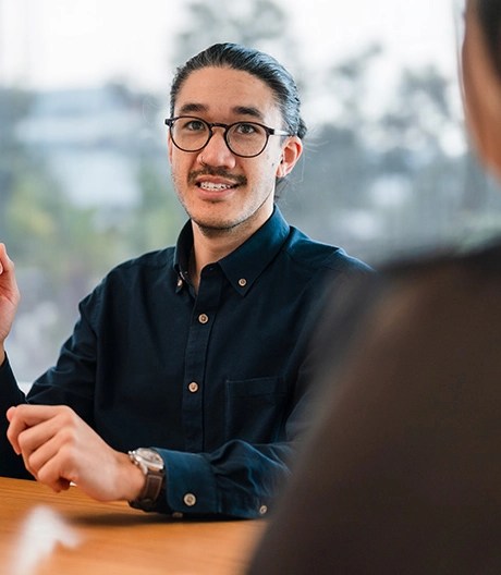 Individual in glasses and a dark shirt speaking across a table to another person who is partially visible, in a professional setting with large windows in the background.