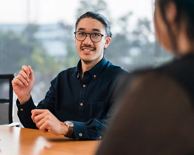 Individual in glasses and a dark shirt speaking across a table to another person who is partially visible, in a professional setting with large windows in the background.