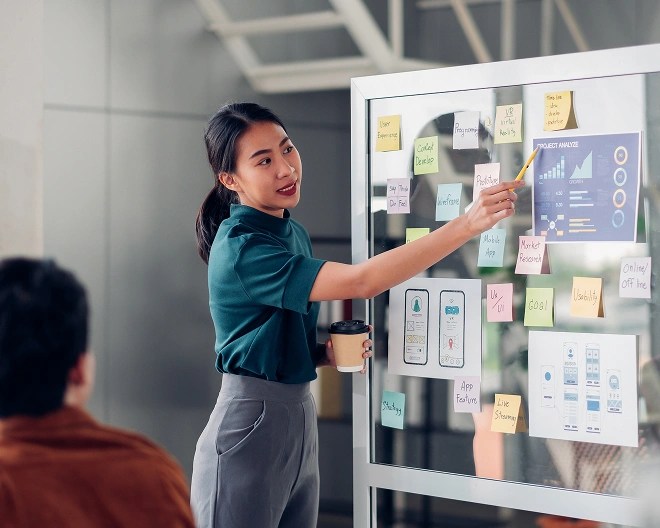Woman holding a coffee cup and pointing to a chart on a glass board covered with sticky notes and printed graphs, in a modern office setting.