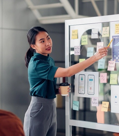 Woman holding a coffee cup and pointing to a chart on a glass board covered with sticky notes and printed graphs, in a modern office setting.