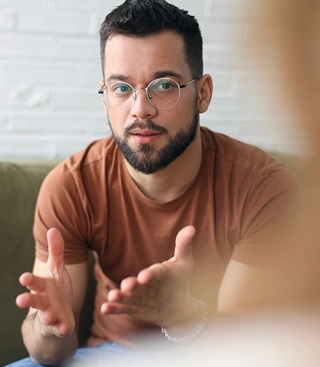 Person in a brown t-shirt and glasses gesturing with both hands while seated on a green couch against a white brick wall, engaged in a discussion with someone in the foreground.