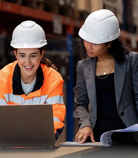 Two individuals in white safety helmets working together in a warehouse, with one wearing a high-visibility jacket and using a laptop, and the other reviewing documents at a table.