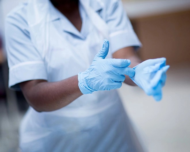 Person in a light medical uniform adjusting blue disposable gloves, in a blurred clinical or hospital setting.
