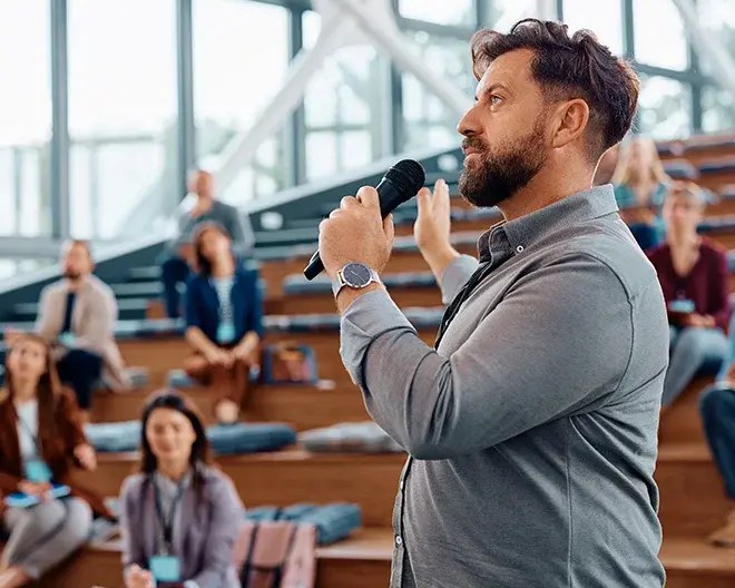 A public speaker wearing a blue shirt talking on a microphone to a group of people in an auditorium.