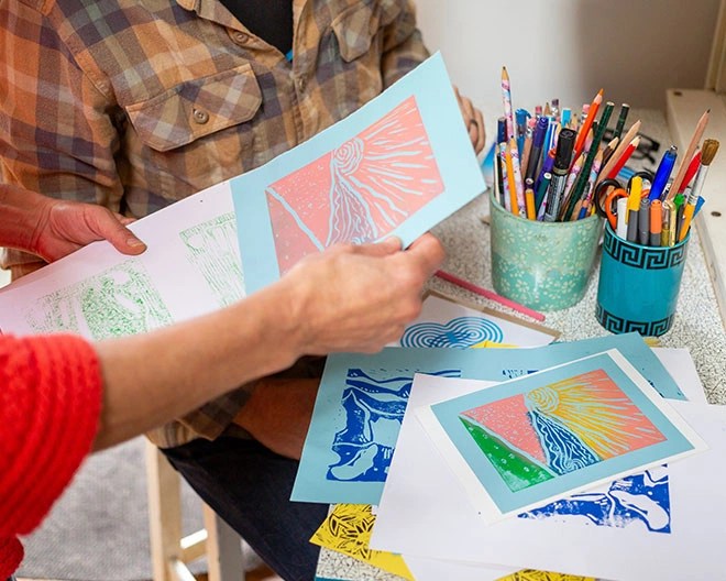 Hands holding paper illustrations with another person in the background and art and pencils on the table