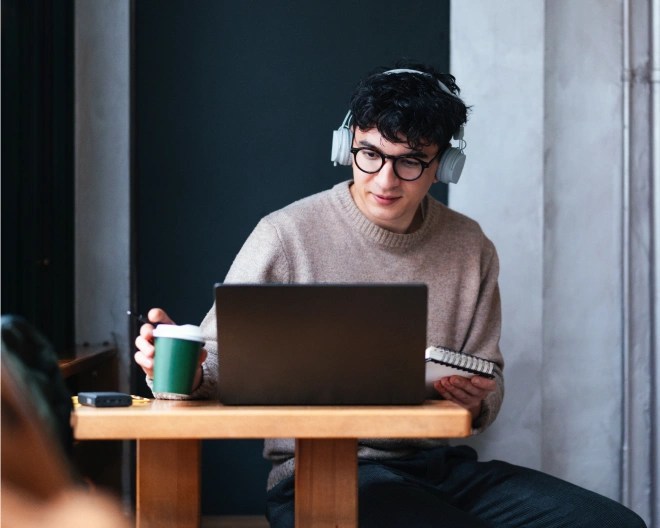 A person wearing wireless headphones picks up their disposable coffee cup while holding a pen with one hand and holds a notepad in another while seated at a desk in front of an open laptop.