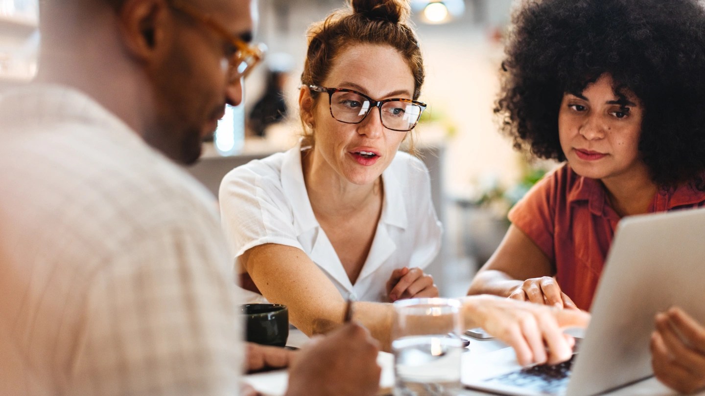 A group of three colleagues sat at a desk looking at a shared laptop, while one of them discusses what is on the laptop.