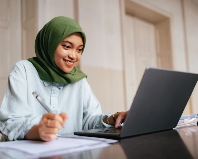 A woman wearing an olive green hijab and a light blue shirt smiles while working on a laptop, holding a pen in one hand.