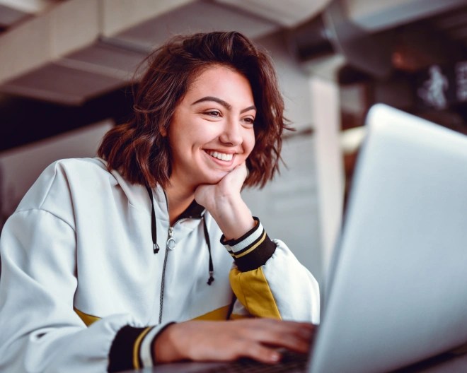 A woman wearing a white sports jacket rests her hand on her chin and smiles while working on a laptop.